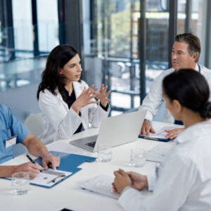 Shot of a group of doctors having a meeting in a modern hospital Grupo de profesionales de la salud discutiendo estrategias en una reunión, con una mujer en bata blanca liderando la conversación frente a una computadora portátil.