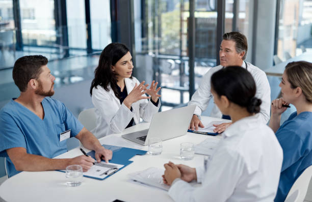 Grupo de profesionales de la salud discutiendo estrategias en una reunión, con una mujer en bata blanca liderando la conversación frente a una computadora portátil.