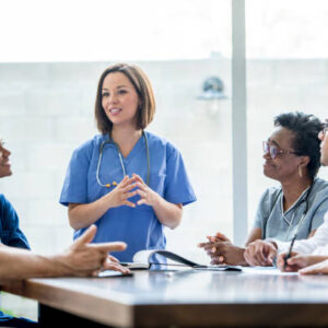 istockphoto-862229772-612×612 Médico en uniforme azul liderando una reunión con un grupo diverso de profesionales de la salud, discutiendo temas importantes en un entorno clínico.
