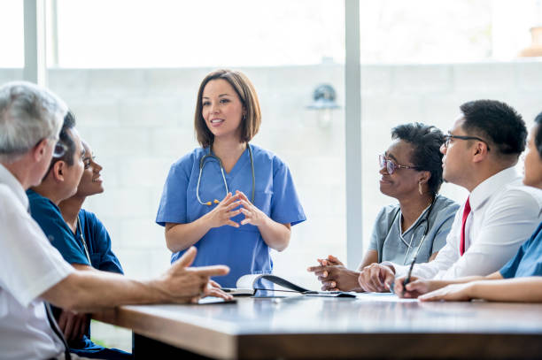 Médico en uniforme azul liderando una reunión con un grupo diverso de profesionales de la salud, discutiendo temas importantes en un entorno clínico.